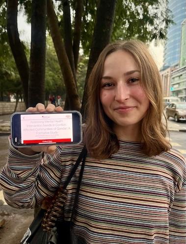 Brooke Erickson holding up her phone that has a slide with the title "Assessing Mental Health Intervention Needs in Conflict-Affected Communities of Gondar: A Formative Study."
