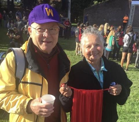 King, wearing a purple Husky hat, and his wife Virginia, who is holding up a red piece of cloth, smile at the camera while standing in a grass area.