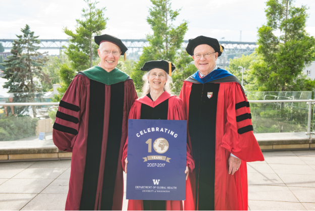 Steve Gloyd, Judy Wasserheit, and King Holmes smile while wearing graduation regalia. Judy holds a sign that says Celebrating 10 Years of DGH, 2007-2017.