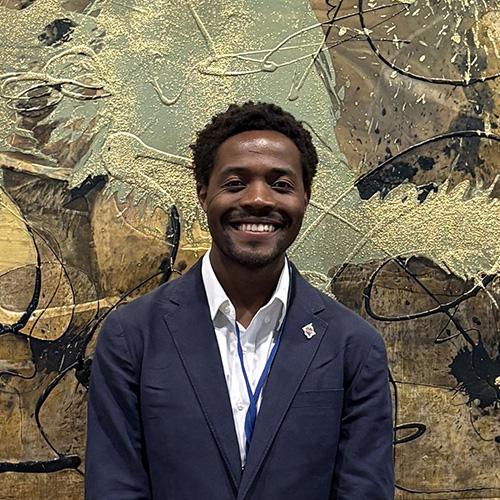 Headshot of Mapenzi Kinege wearing professional attire in front of a painting.